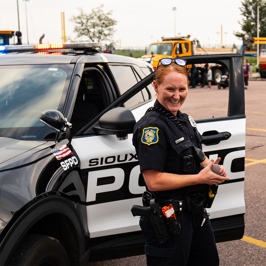 Police officer at a community event standing beside a Sioux Falls Police Department patrol car, smiling, with emergency lights activated in the background.